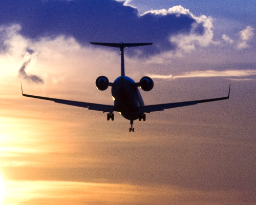 A small jet aircraft seen from behind as it descends toward a runway at sunset, silhouetted against a sky with golden and blue clouds.