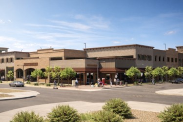 Large tan commercial building at a street corner with sidewalks, small trees, parked cars, and a few pedestrians under a blue sky.