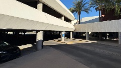 Covered drive-through area beneath a multi-level public parking garage, with vehicle lanes, structural columns, and pedestrian access in daylight.