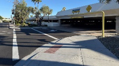 Pedestrian crosswalk and sidewalk ramp leading to a public parking garage entrance, with clearance signage, trees, and desert landscaping in daylight.