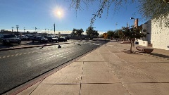 Wide sidewalk along a street with a marked crosswalk, parked cars, trees, and a public building in bright sunlight.