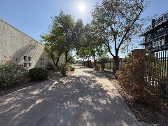 Sunlit walkway curving between a textured light‑colored building and a tall metal fence, surrounded by desert plants, trees, and landscaped gravel.