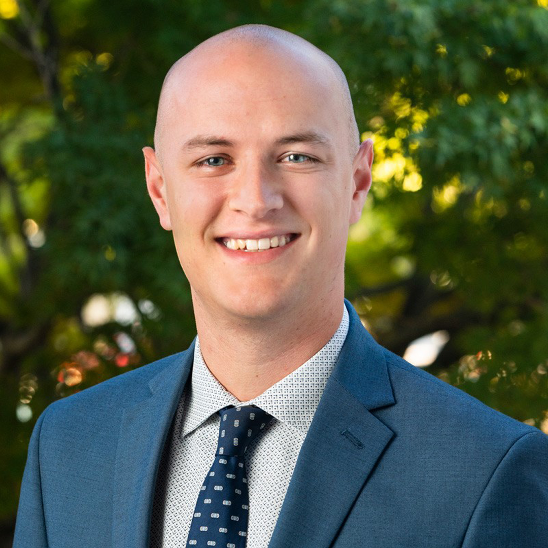 Professional headshot of a man in a blue suit and patterned tie, smiling outdoors in front of green trees.