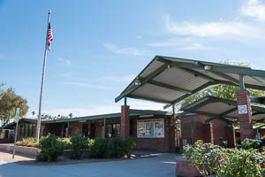 Brick neighborhood center with green awnings, landscaping, and a nearby flagpole under blue sky.