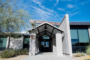 Modern community center entrance with tall pillars, glass windows, and desert landscaping beneath blue sky.