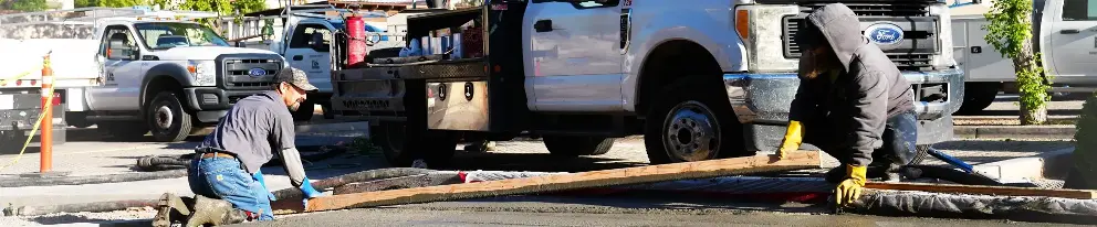 Two workers smoothing wet concrete near parked trucks on a construction site.