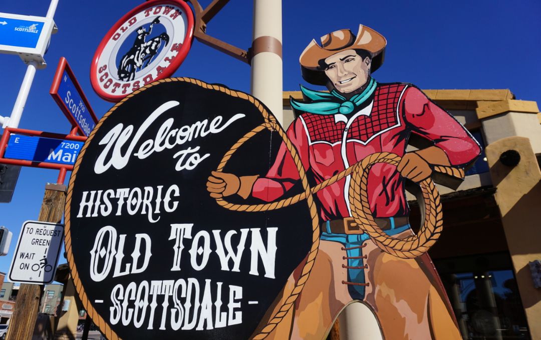 Illustrated cutout of a smiling cowboy holding a lasso beside a black sign reading Welcome to Historic Old Town Scottsdale, with an Old Town Scottsdale logo above and street signs for Scottsdale and Main.