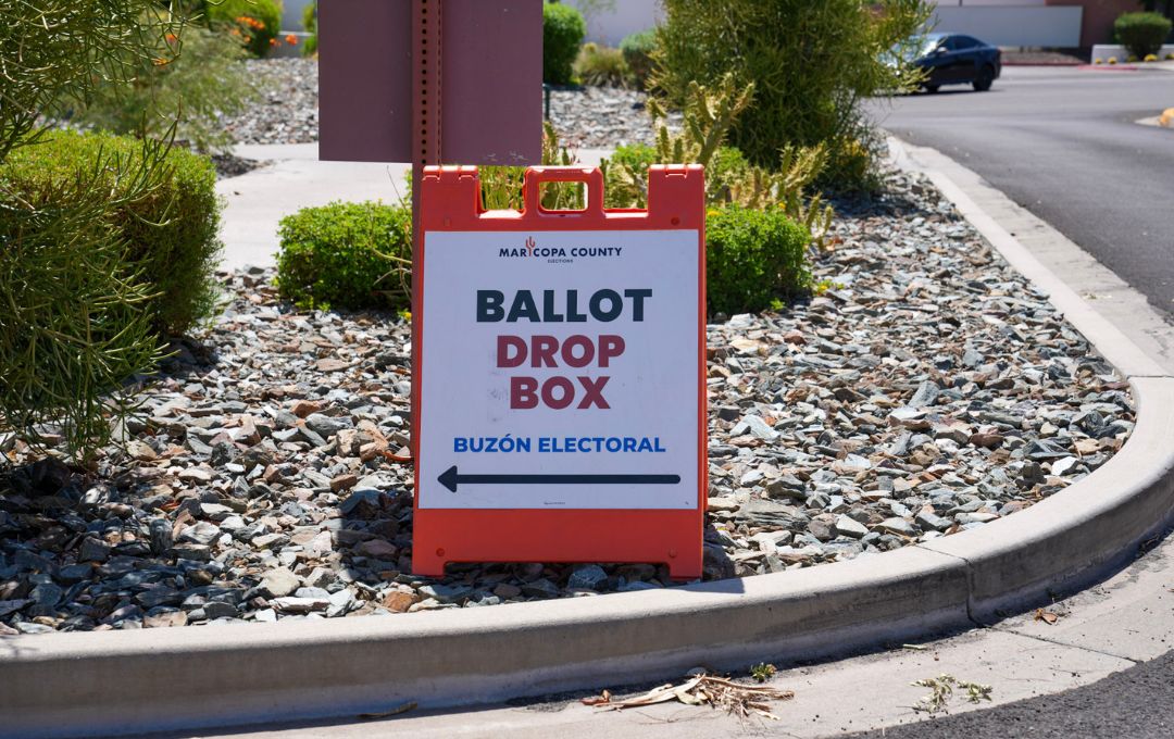 Maricopa County Elections sign in a rock landscaping bed reading Ballot Drop Box and Buzón Electoral, with an arrow pointing left toward the drop-off location.