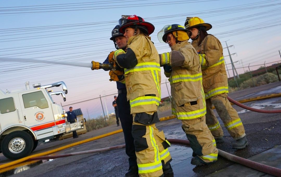 Four people in firefighter gear hold a high-pressure hose, with a fire truck in the background.