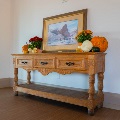 A carved wood console table decorated with pumpkins, gourds and mums beneath a framed western painting of riders near a red rock formation.
