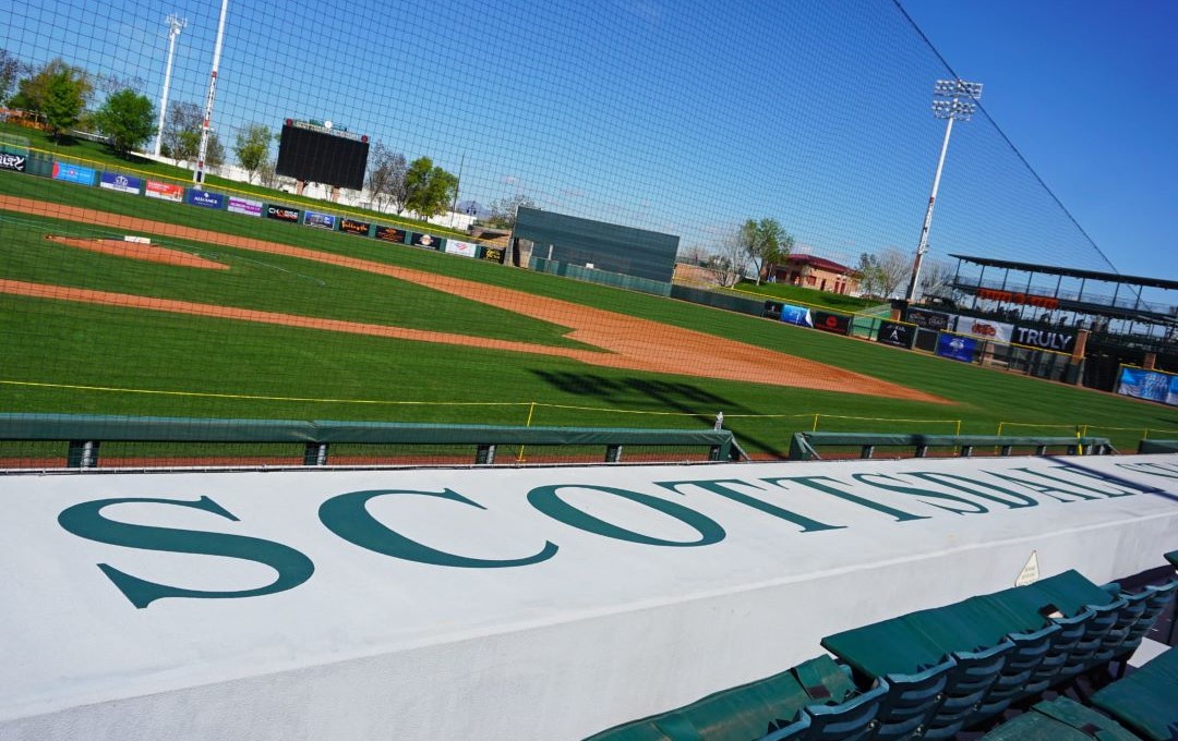 View of a baseball field through protective netting, overlooking a white dugout roof with "SCOTTSDALE" painted in green.