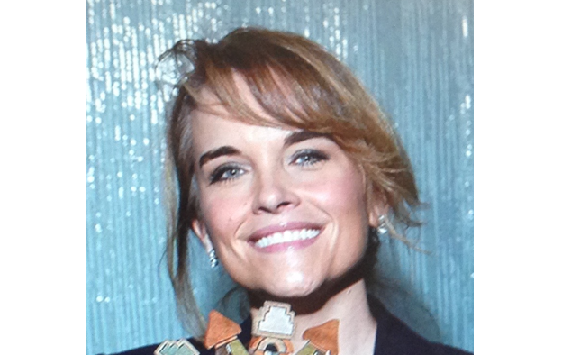 A smiling woman with light brown hair, bangs, and blue eyes against a shimmering silver background.