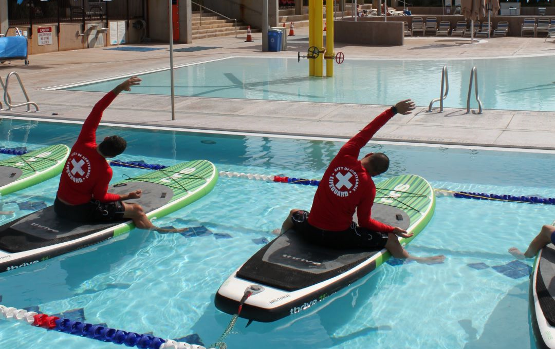 Two lifeguards practicing paddleboard yoga stretches in an outdoor swimming pool.
