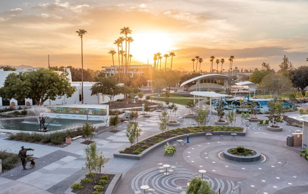 A sunset overlooks a modern plaza with palm trees, a water fountain, sculptures, and a large canopy.