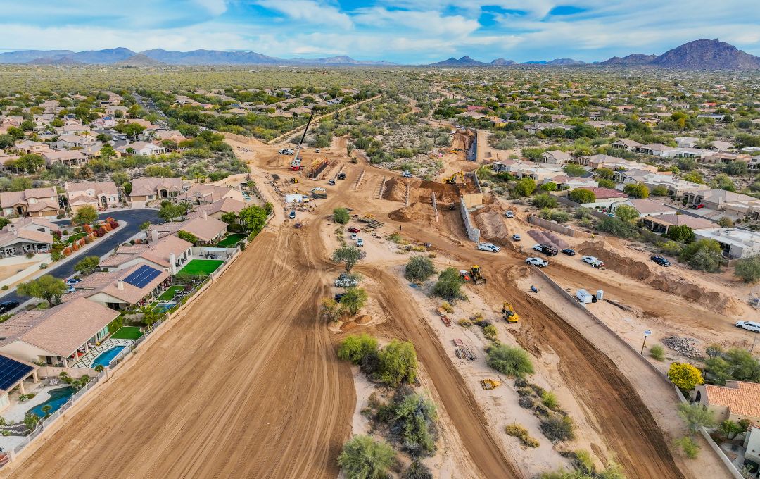 Aerial view of the Rawhide Wash Phase 1 construction site, a wide graded dirt corridor with cranes, excavators, and trucks cutting between two residential neighborhoods, with desert and mountains in the distance.