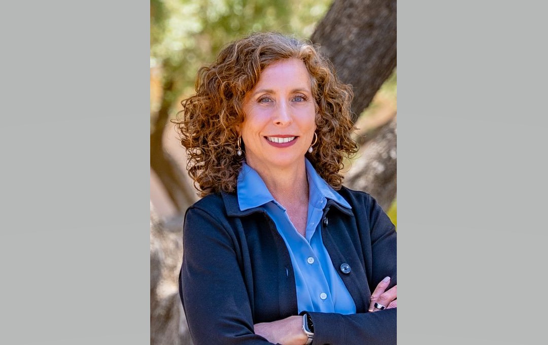A professional portrait of a smiling woman with curly brown hair, wearing a blue shirt and black jacket.