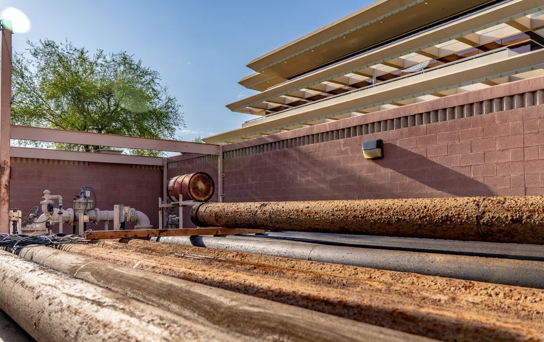Large rusted industrial water pipes lying outdoors near a brick wall and modern building.