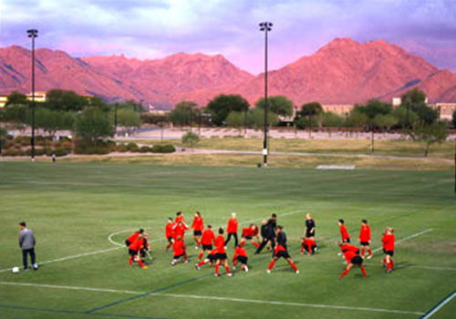 Group practicing on a large green sports field with tall light poles, set against a backdrop of rugged mountains under a colorful sky.