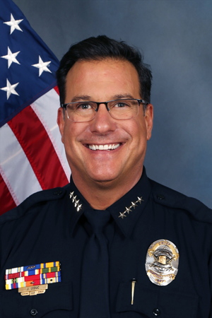 Formal portrait of a smiling police chief in dress uniform, standing in front of an American flag backdrop.