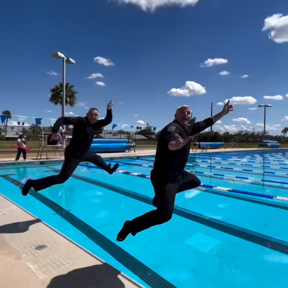 Twoindividualsindarkuniformsjumpingintoabrightblueoutdoorpoolcapturedmidairunderasunnyskywithscatteredclouds