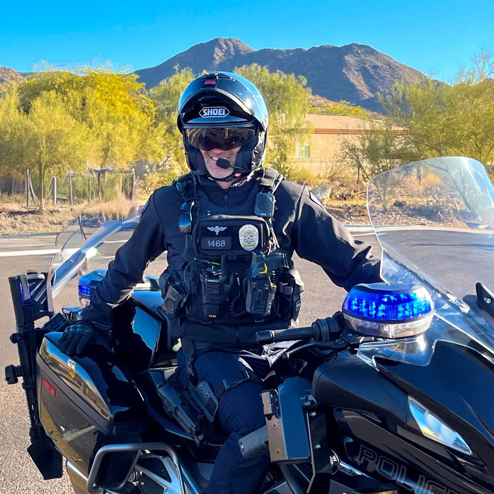 Asmilingpolicemotorcycleofficerinahelmetsitsonhisbikewithdesertmountainsbehindhim