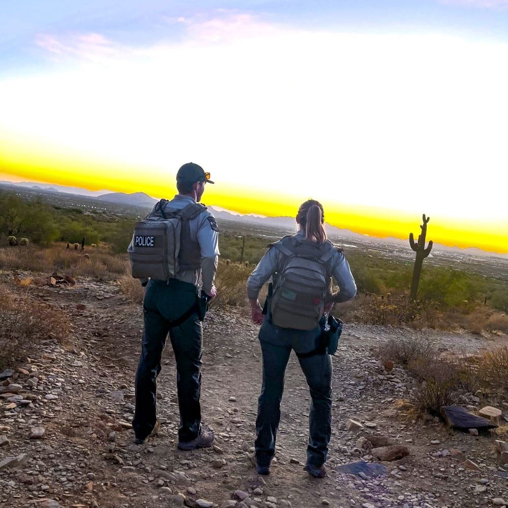 Twopoliceofficerswithbackpackslookoutoveradesertlandscapeatsunset