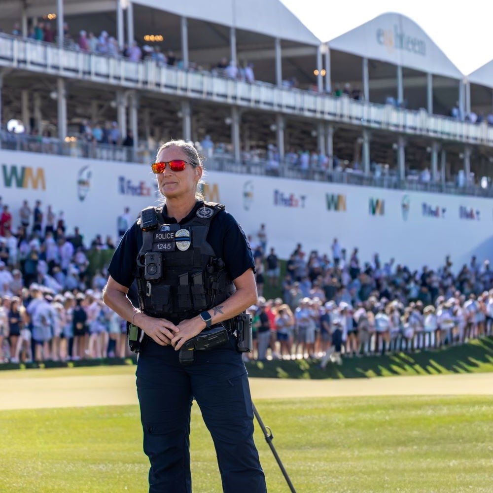 Apoliceofficerinsunglassesstandsonagolfcoursewithlargecrowdsandtentsbehindher