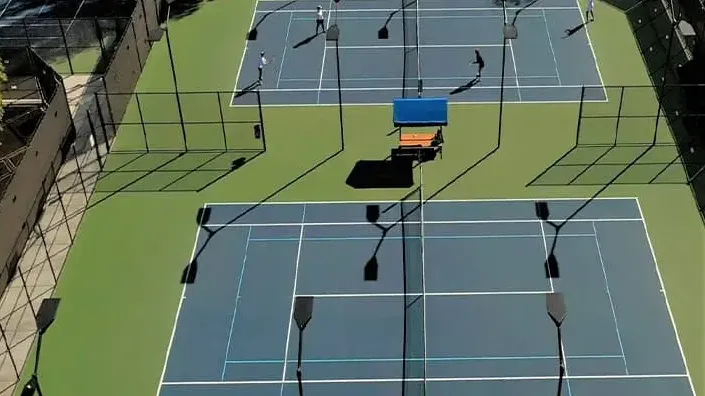 Players practicing on multiple blue-and-green outdoor tennis courts surrounded by tall fencing.