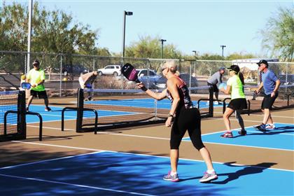 Six adults playing pickleball on adjacent blue outdoor courts under bright, sunny desert skies.