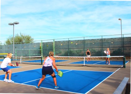 Four women playing pickleball on bright blue outdoor courts surrounded by fencing and lights.