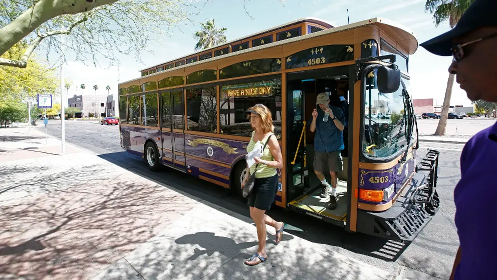 A trolley-style bus with "Have a Nice Day" on its side panel is at a stop. People are boarding and alighting, with a woman in casual attire stepping off. A man in purple watches from the foreground.