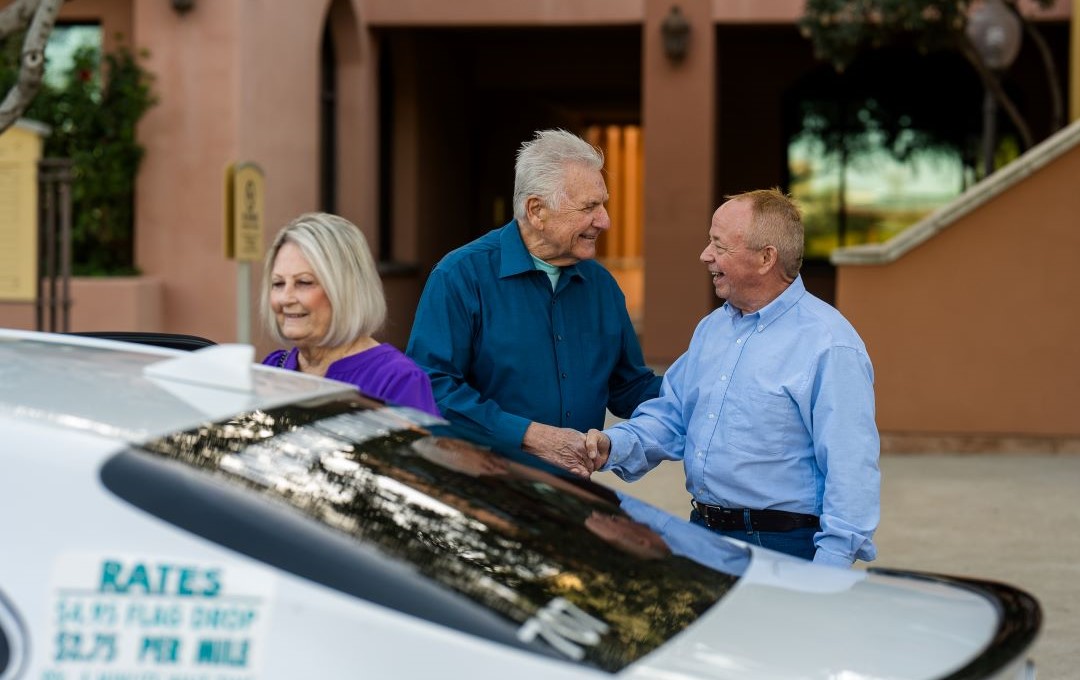 A smiling elderly man in a blue shirt shakes hands with another man, with a woman beside him, next to a car.