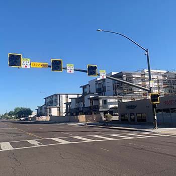 Pedestrian crosswalk with overhead flashing signals spanning the road, near modern buildings under a clear blue sky.