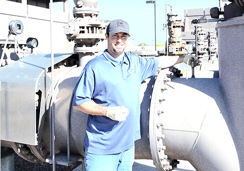 Individual wearing a cap and work gloves standing beside large industrial pipes and valves outdoors, resting one hand on a bolted connection.