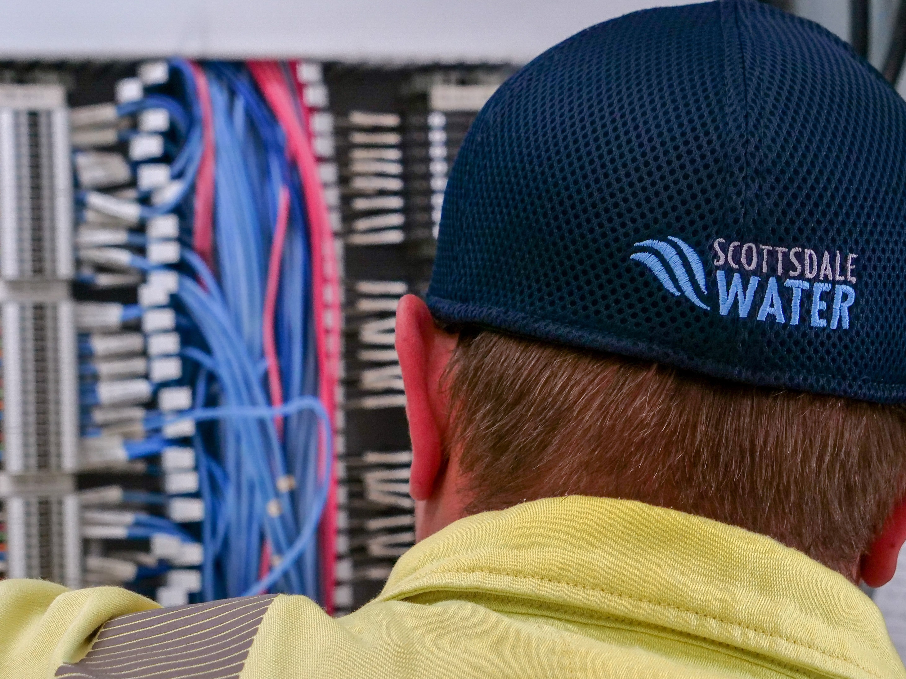 Individual wearing a Scottsdale Water cap standing in front of a control panel filled with organized bundles of blue and red wires.