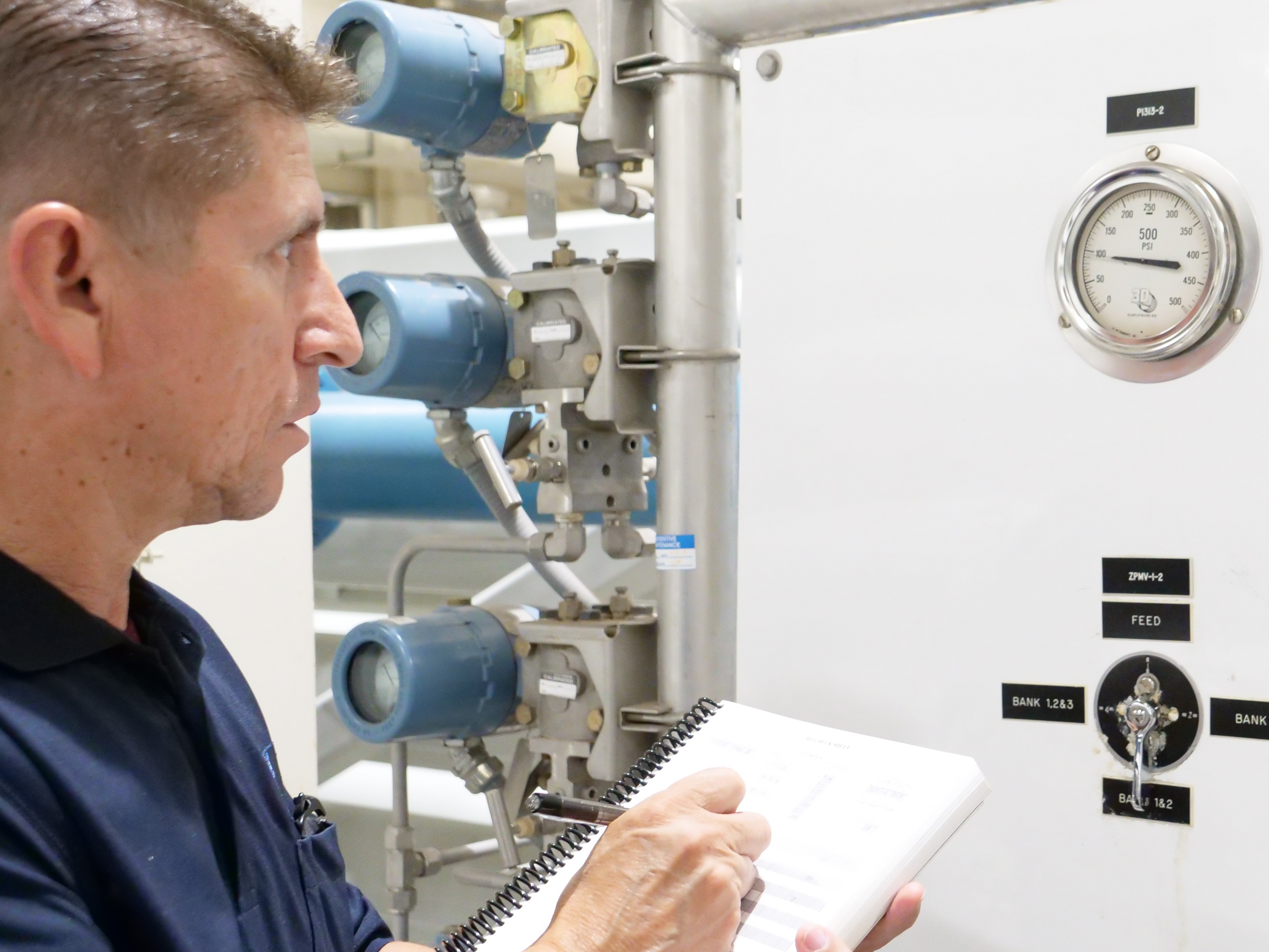 Individual examining gauges and equipment in an industrial facility while writing notes in a spiral notebook.