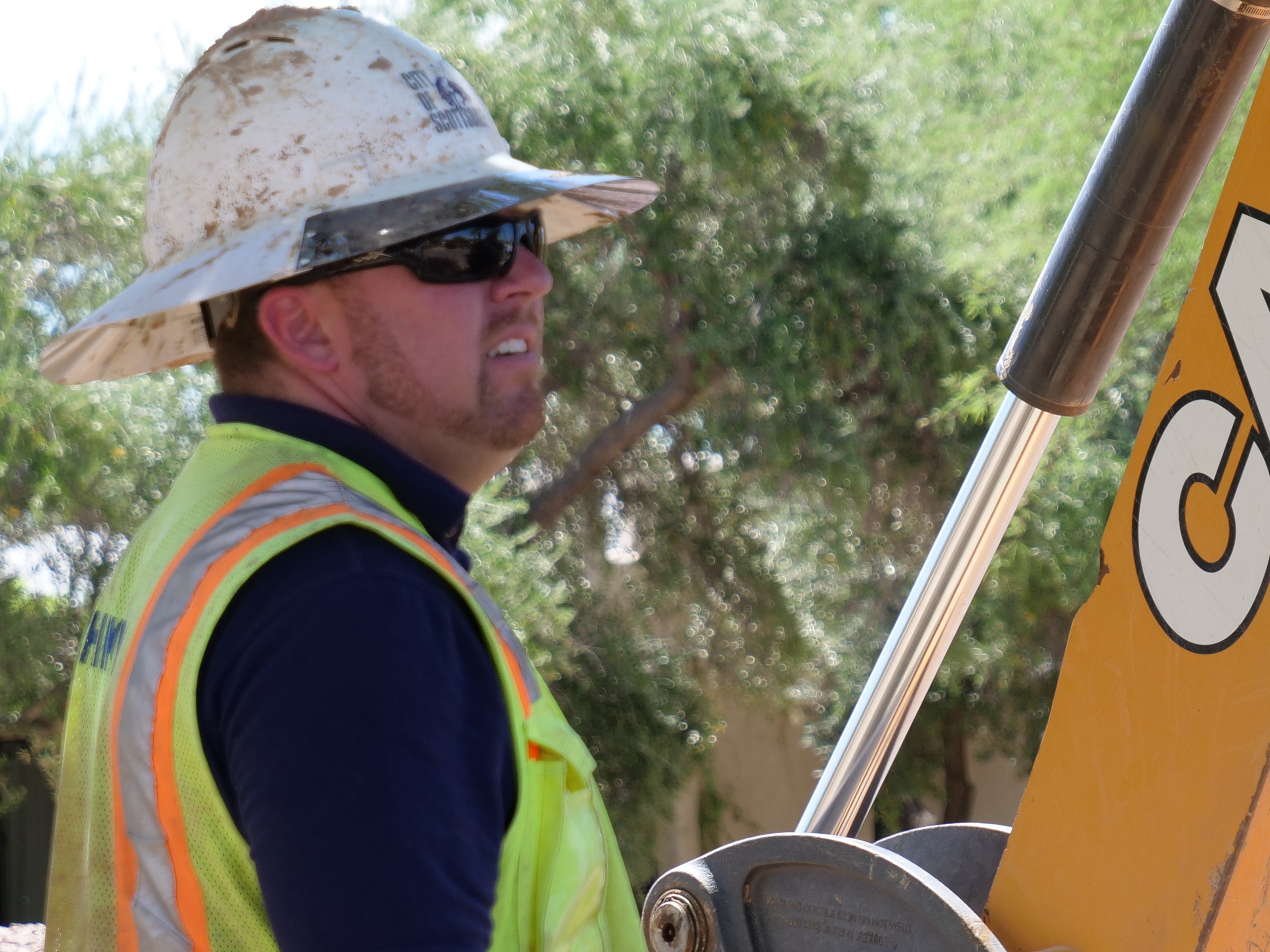 Construction worker wearing safety gear, including a hard hat and reflective vest, standing beside heavy equipment outdoors.