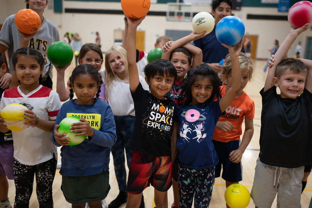 Group of young children smiling and holding colorful balls during an indoor sports activity.