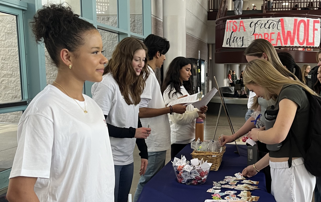 Students at table handing out materials and treats during school event in busy lobby.